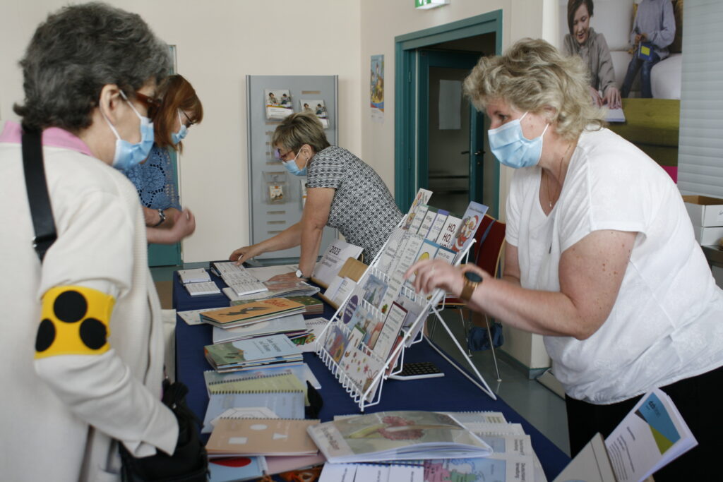 Ein Verkaufstisch mit vielen Büchern, Karten und Reliefs, dahinter zwei Frauen mit Maske, davor zwei Besucherinnen, die sich die Produkte erklären lassen