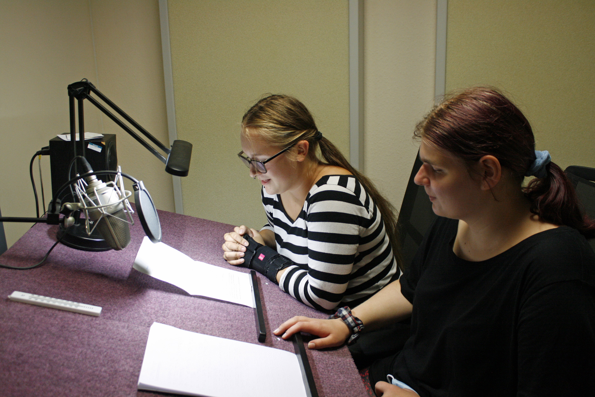 Zwei weibliche Jugendliche sitzend vor einem Mikrofon und ihrem Text, die eine mit Brille und geringeltem Sweatshirt spricht ins Mikrofon