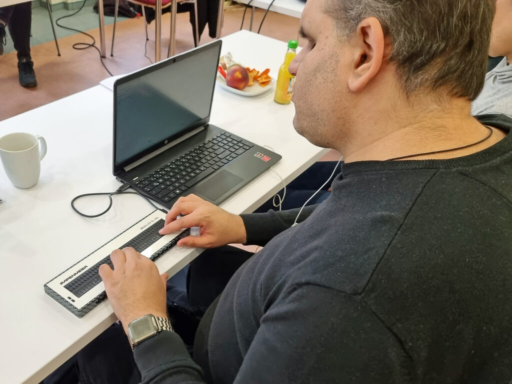 Ein Teilnehmer am Tisch sitzend liest mithilfe der Braillezeile, rechts Latop