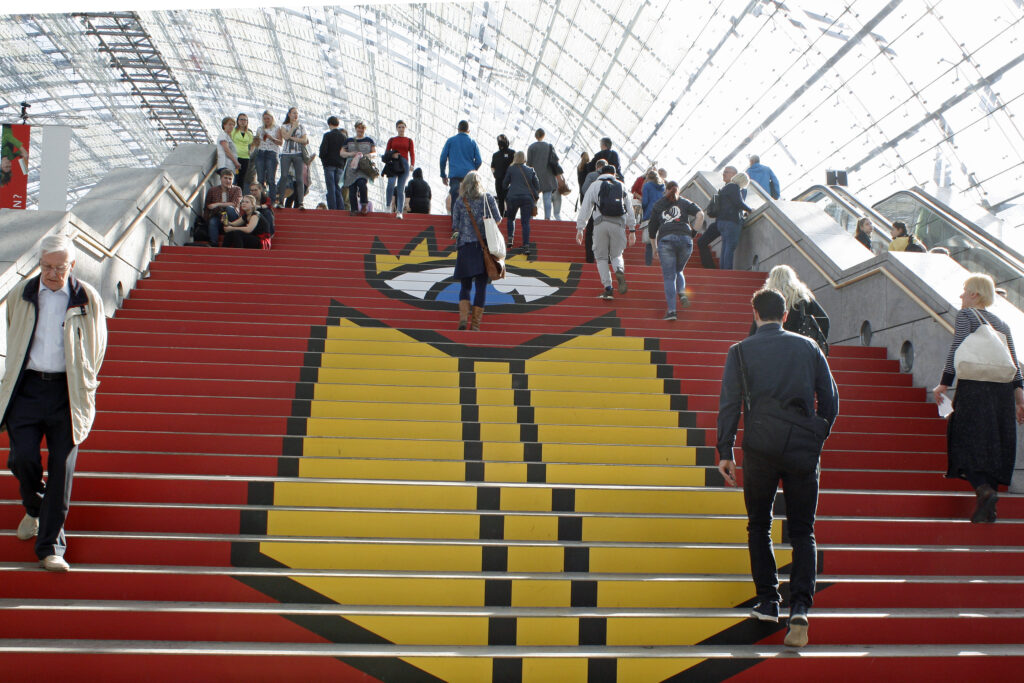 Große rote Treppe mit Buchmesse-Logo (Buch und Auge) in der Glashalle. Menschen laufen auf der Treppe.