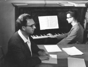 Im Vordergrund ein Mann mit Brille, der Braillenoten liest, im Hintergrund ein Mann mit Brille am Klavier
