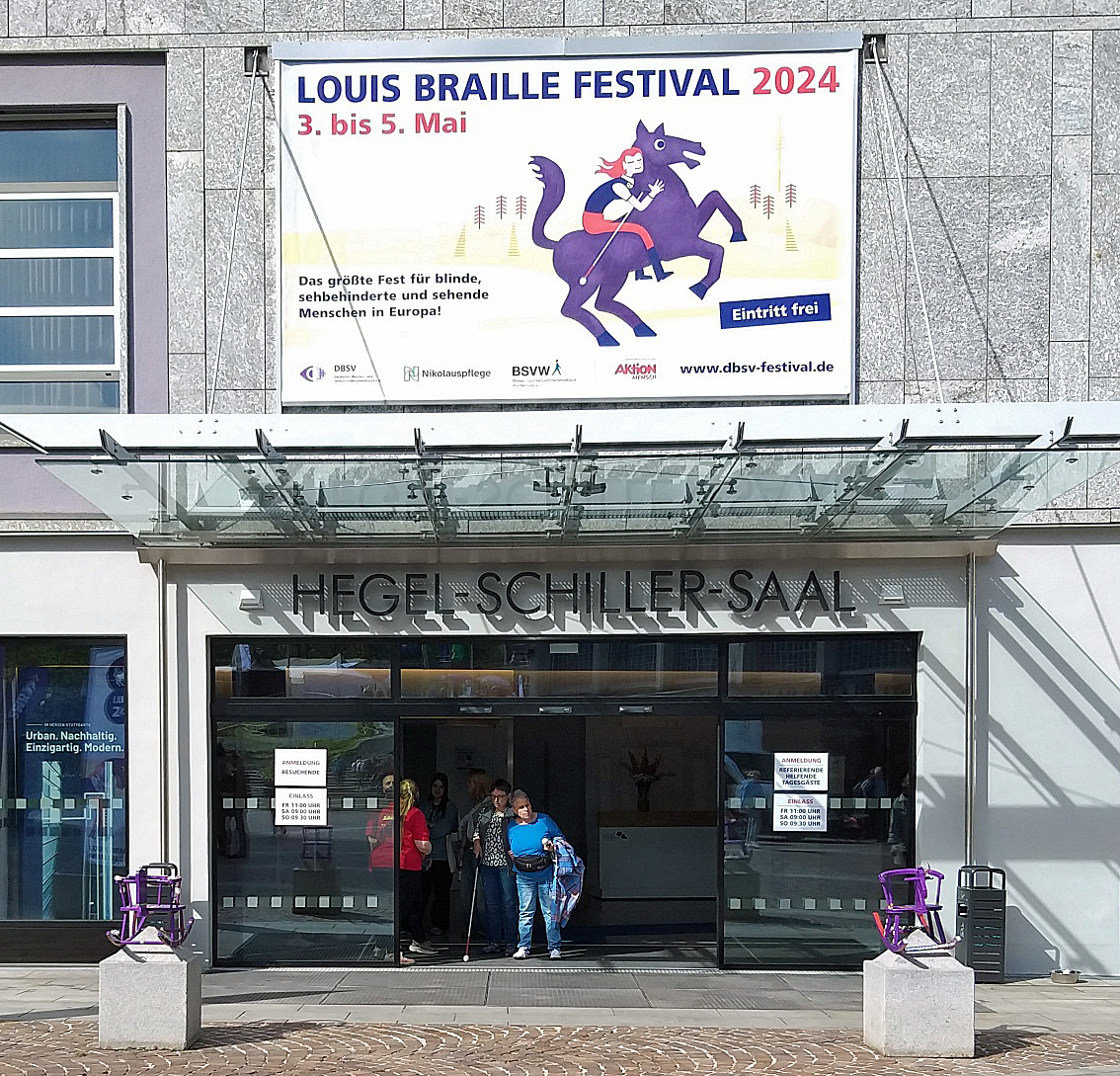 Eine großer Eingang mit Überdachung, davor links und rechts zwei lilafarbene Schaukelpferde, darüber ein großes Plakat "Louis Braille Festival 2024 mit lila Rössle und Reiterin