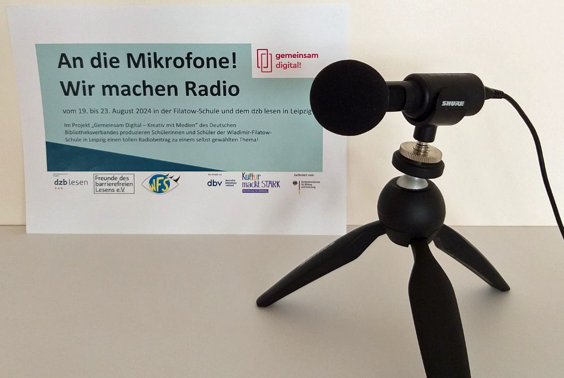 Ein Mikrofon im Vordergrund, dahinter ein kleines Plakat "An die Mikrofone! Wir machen Radio"