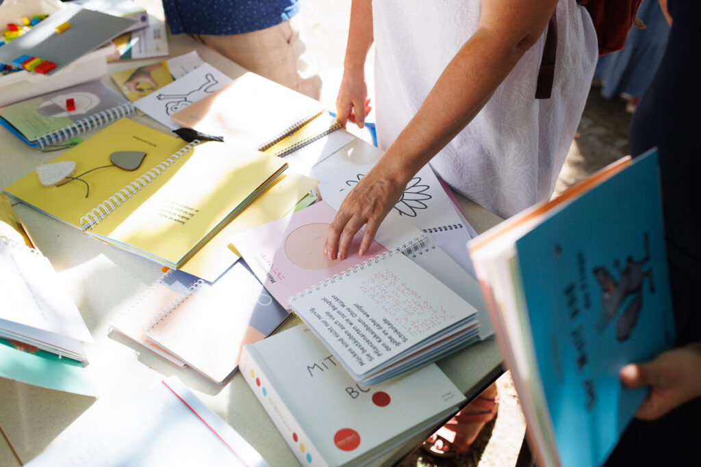 Viele aufgeschlagene tastbare Kinderbücher liegen auf einem Tisch. Eine Hand ertastet eine Illustration