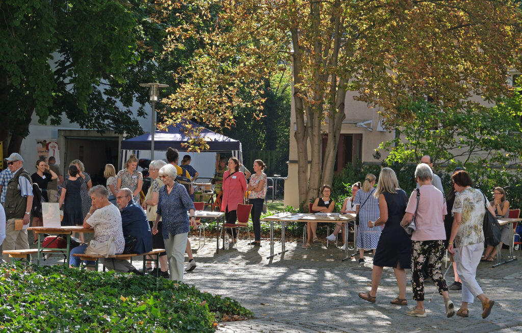 Links sitzen einige Menschen auf Bänken, in der Mitte stehen zwei Frauen neben einem Tisch, daneben sitzt zwei Frauen am Tisch, ganz rechts laufen mehrere Personen am Stand vorbei, im Hintergrund steht ein Baum vor einem Haus. 