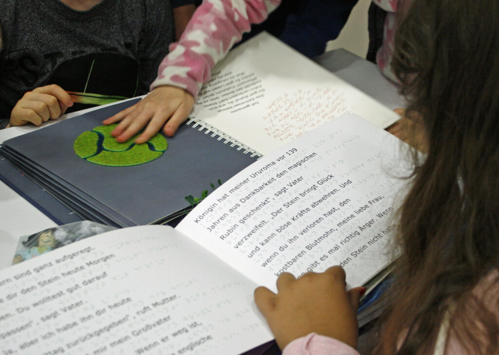 zwei aufgeschlagene Bücher in Braille- und Schwarzschrift werden von zwei Händen ertastet