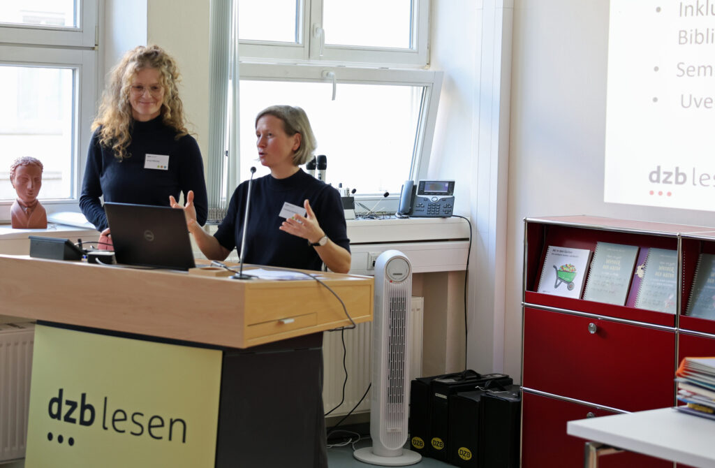 Zwei Frauen am Rednerpult: Die linke mit langen Haaren lächelt, die rechte mit kurzen Haaren spricht zum Publikum.
