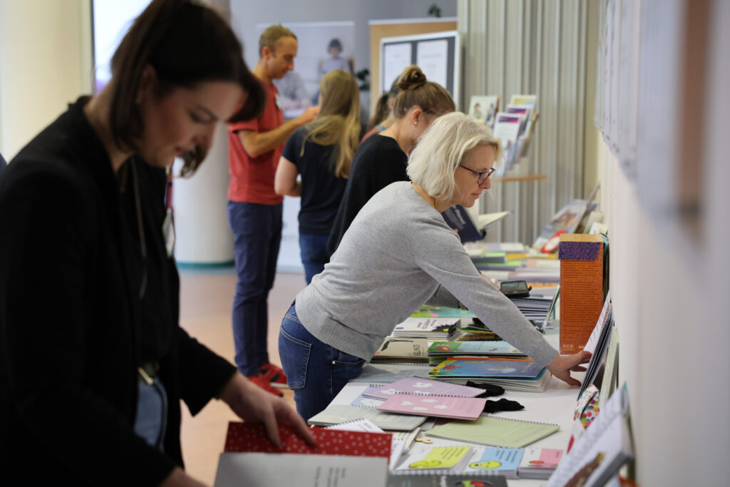Mehrer Personen stehen vor Büchertischen und schauen sich taktile Bilderbücher an. Im Hintergrund drei Personen im Kreis und unterhalten sich.