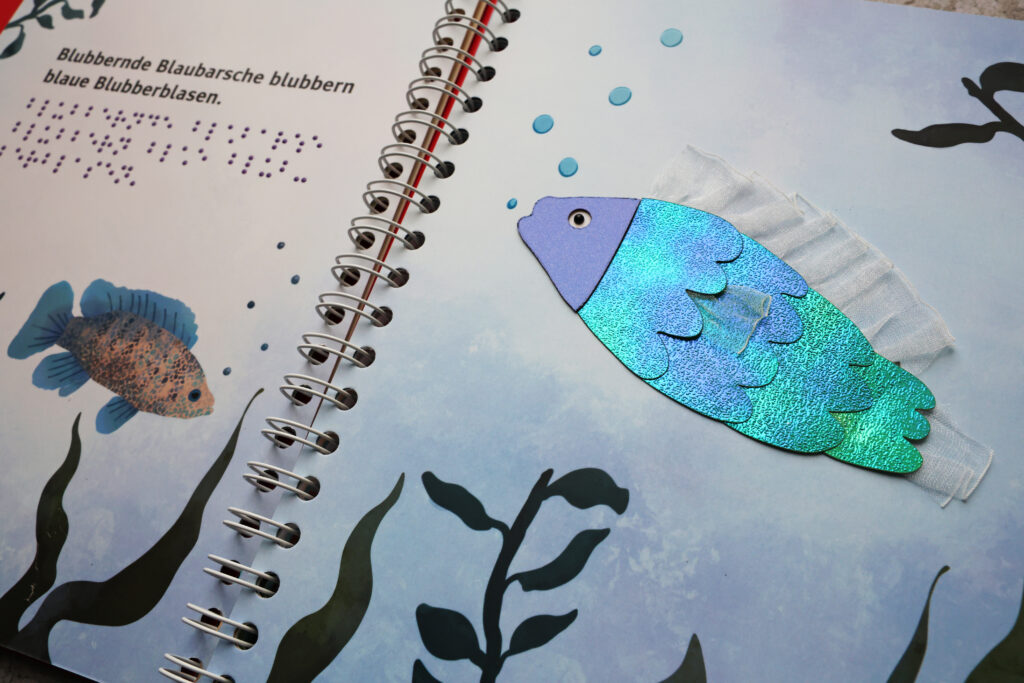 Ein aufgeschlagenes Ringbuch: die linke Seite mit Braille- und Großschrift, rechte Seite mit tastbarer Illustration (Fisch).