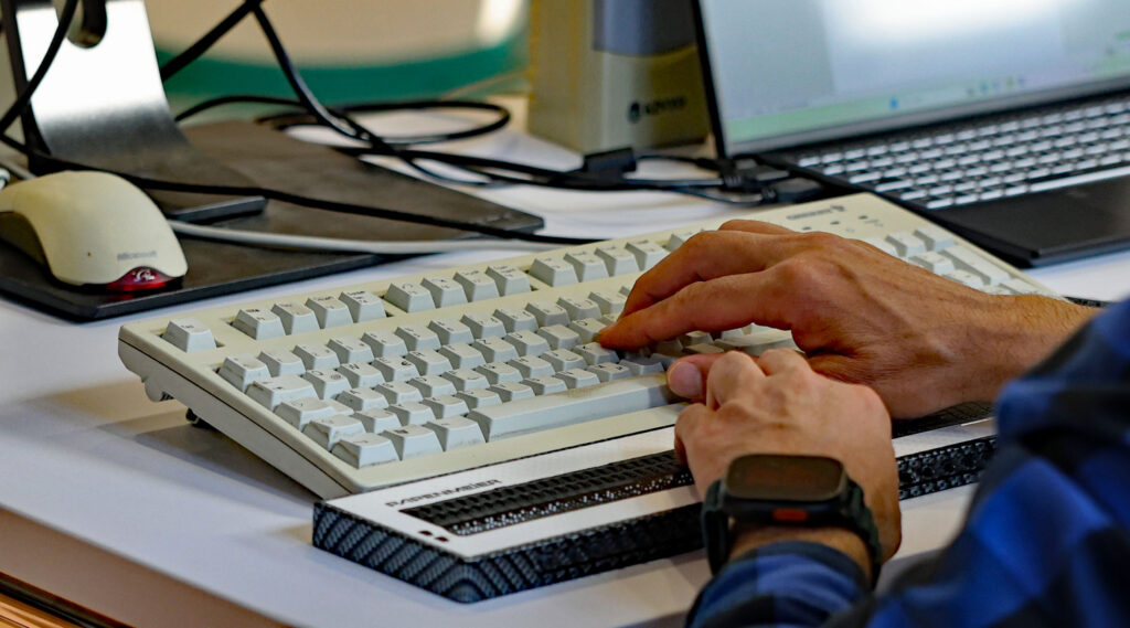 Zwei Hände auf der Tastatur eines Computers und der Braillezeile, dahinter angeschnitten ein Laptop und eine Mouse