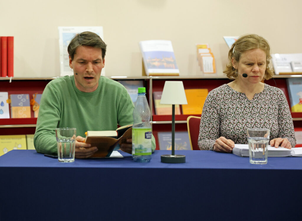 Zwei Personen an einem Tisch: links ein Mann im grünen Pullover hält ein Buch in den Händen und liest, rechts eine Frau mit schulterlangem Haar und Mikro hat vor sich ein Braillebuch liegen. Hinter ihnen steht ein Sideboard mit Büchern