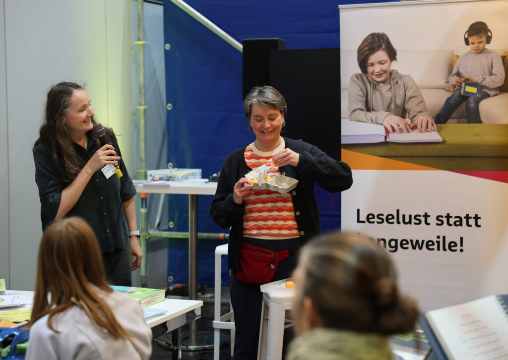 Zwei Frauen vor jungem Publikum: links die eine mit langen Haaren hält ein Mikrophon in den Händen und spricht, rechts eine Frau mit kurzen Haaren hält eine Eierpackung mit drei Tennisbällen in den Händen. Rechts ein Plakat mit der Aufschrift "Leselust statt Langeweile"