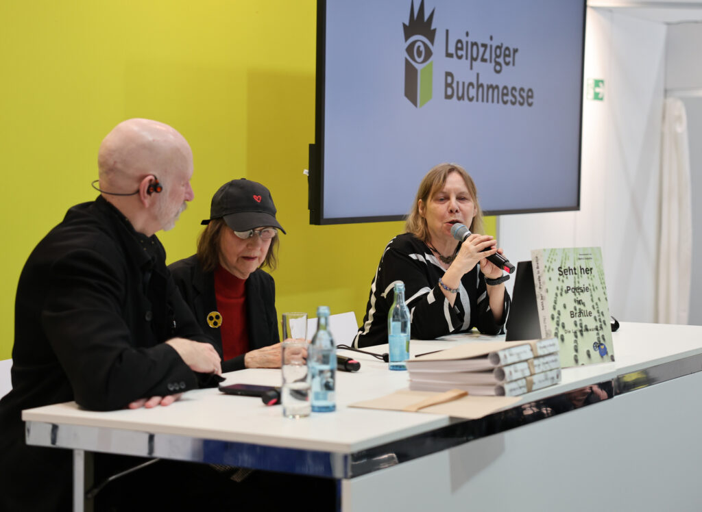 Drei Personen sitzen vor einem Tisch in einem Forum. Rechts spricht eine Frau ins Mikrophon, neben ihr eine Frau mit Cape und neben ihr, ganz links ein Mann mit Glatze, auf dem Tisch stehen Getränke und liegen Bücher.
