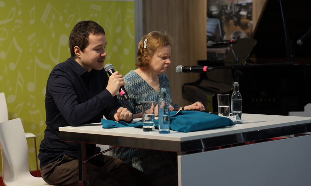 Zwei Personen am Tisch: links ein junger Mann mit Oberlippenbart spricht ins Mikrophon, rechts eine Frau mit brünettem, schulterlangen Haar, vor ihrem Mund ein Mikrophon. Auf dem Tisch stehen zwei Gläser und Flaschen mit Wasser