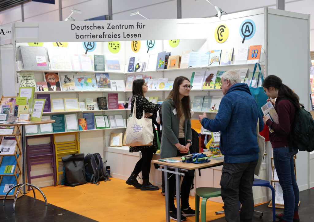 der Stand des dzb lesen mit vielen Regalen voller Bücher, eine Besucherin schaut sich ein Buch aus dem Regal an, im Vordergrund erklärt eine junge Frau mit langen Haaren einem Besucher etwas.