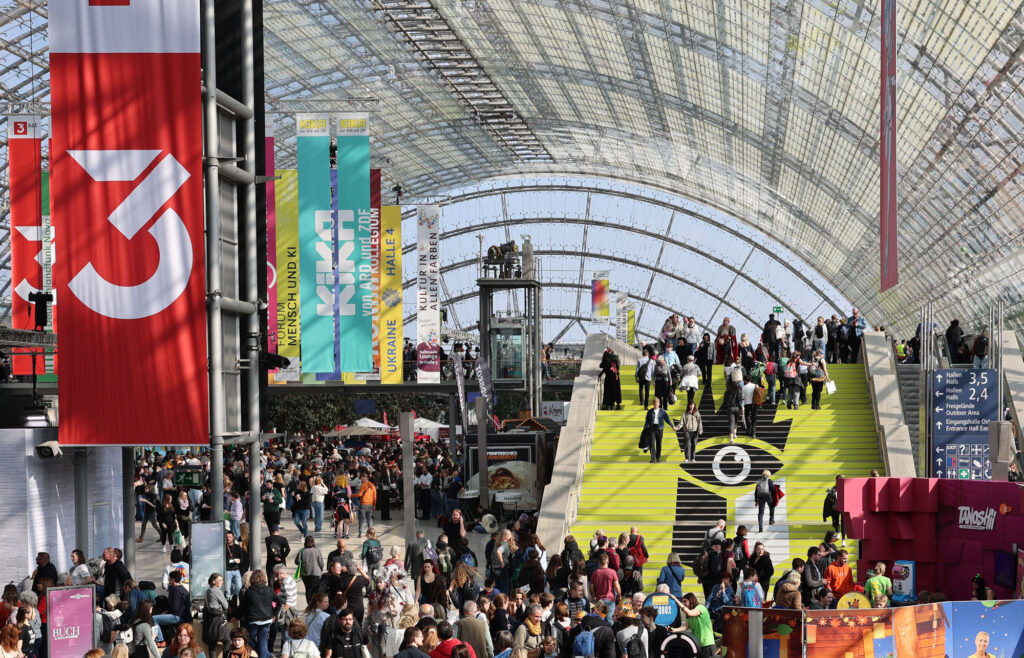 Ein Glashalle mit vielen Menschen, die durch die Halle strömen, rechts eine große Treppe mit Messe-Logo, Links hängen viele Werbeplakate