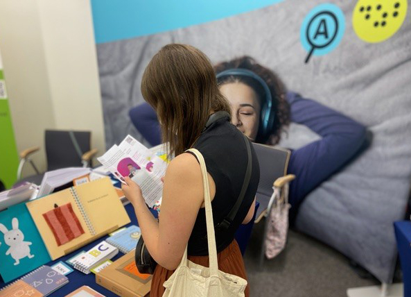 Eine Messebesucherin steht vor einem Tisch, auf dem mehrere taktile Kinderbücher liegen. Sie liest in einer Broschüre, die sie in den Händen hält.